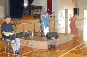 平和について語りエンディングで「童神」を熱唱した古謝さん（中）。キーボード演奏の佐原さん（左）と歌に振り付けたハワイアンフラを踊る森本さん（右）＝27日、久松中学体育館