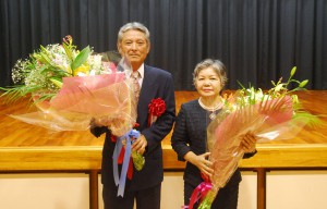 花束を手に参加者に感謝する佐藤さん夫妻＝23日、富名腰公民館