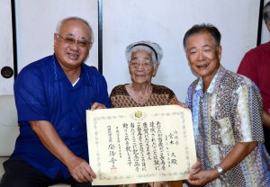 総理大臣表彰を手に笑顔の宮本久さん（中央）。左は下地市長、右は宮本さんの長男の寛忠さん＝平良下里