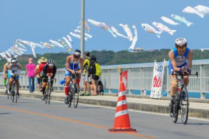 来間大橋に掲げられた初夏を思わせる鯉のぼりの下をバイクで疾走する選手たち