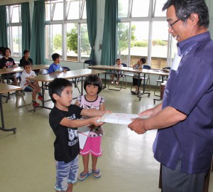 狩俣会長（右）から学用品を受け取る児童＝１１日、上区自治会公民館