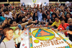 大会ロゴマークをかたどったケーキを囲んで乾杯する選手たち。大会当日の健闘を誓い合った＝１７日、市総合体育館