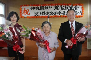 花束を贈呈され祝福を受ける伊佐照雄さん（右）、中央は母親のツルさん、左は妻の美恵子さん＝５月３１日、大神島離島振興コミュニティーセンター