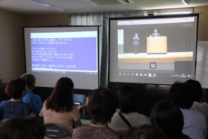 浦添市てだこホールでの片岡市長の基調講演を生中継するサテライト上映。左側は聴覚障がい者に対応した同字幕システムのスクリーン＝８日、介護センター・スマイル