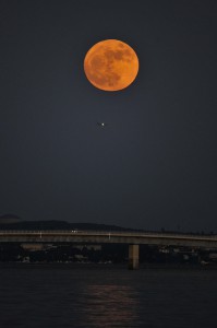 伊良部大橋（下）にかかるスーパームーン。月の下の光の点はジェット旅客機＝１４日午後６時５分すぎ、伊良部島から（金子進撮影）