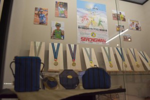 展示されているトライアスロンメメダル帯と（手前左から）ひも付き口ポーチ、お守り、コースター、がま口ポーチ＝１５日、市伝統工芸品センター