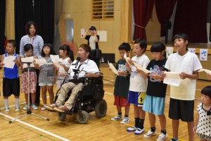 車いすの謝花さんと合唱する児童たち＝20日、狩俣小学校体育館