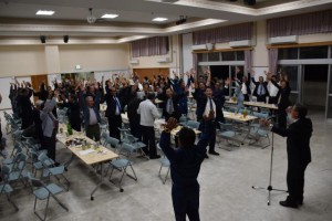 池城三千雄教育長の掛け声に合わせ、万歳三唱する参加者ら＝６日、村コミュニティー施設